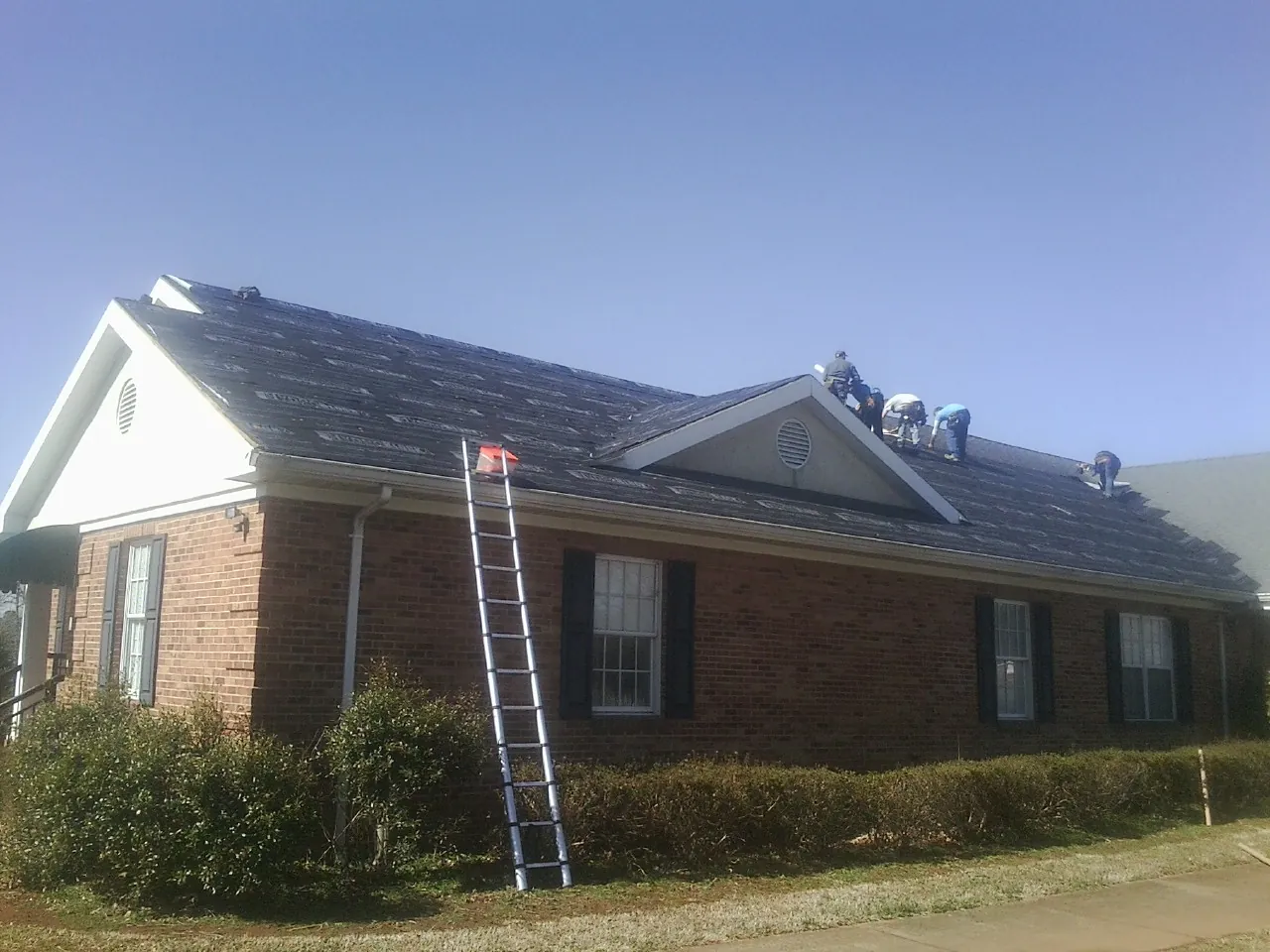 Crew tearing shingles off a brick ranch roof with a ladder set — Emergency Roof Repair work by our crew in Remlap