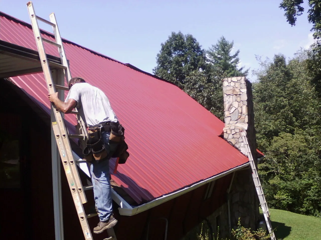 Roofer on a ladder beside a red metal roof and stone chimney — Soffit & Fascia Repair by Roofing Brothers in Remlap